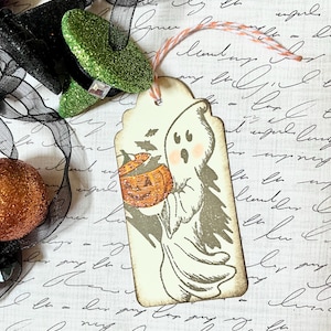 May include: A Halloween-themed gift tag featuring a ghost holding a carved pumpkin. The tag has a scalloped edge and is tied with orange and white twine. The ghost is gray with pink cheeks, and the pumpkin is orange. A black bow and glittery green witch shoe are in the background.