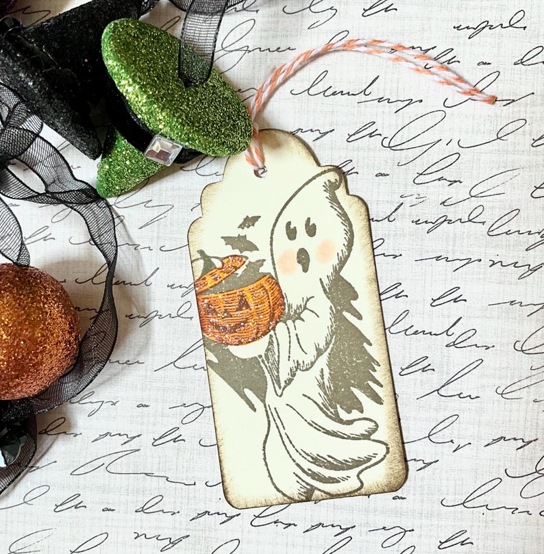 May include: A Halloween-themed gift tag featuring a ghost holding a carved pumpkin. The tag has a scalloped edge and is tied with orange and white twine. The ghost is gray with pink cheeks, and the pumpkin is orange. A black bow and glittery green witch shoe are in the background.