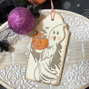 May include: A Halloween-themed tag featuring a friendly ghost holding a glittery orange pumpkin. The tag has a distressed, vintage look with a string for hanging. The ghost has rosy cheeks and is surrounded by bats. Decorative purple and orange glitter balls are in the background.