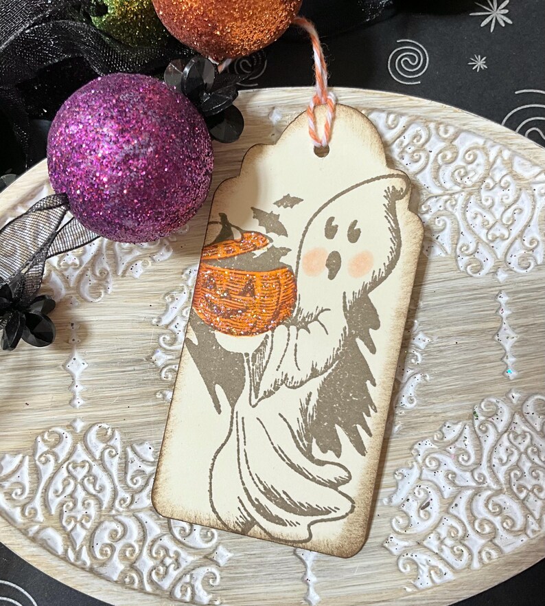 May include: A Halloween-themed tag featuring a friendly ghost holding a glittery orange pumpkin. The tag has a distressed, vintage look with a string for hanging. The ghost has rosy cheeks and is surrounded by bats. Decorative purple and orange glitter balls are in the background.