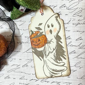 May include: A Halloween-themed gift tag featuring a friendly ghost holding a jack-o'-lantern. The tag has a distressed, vintage look with a string for hanging. The ghost has rosy cheeks and is surrounded by bats. The tag is cream-colored with a brown border.