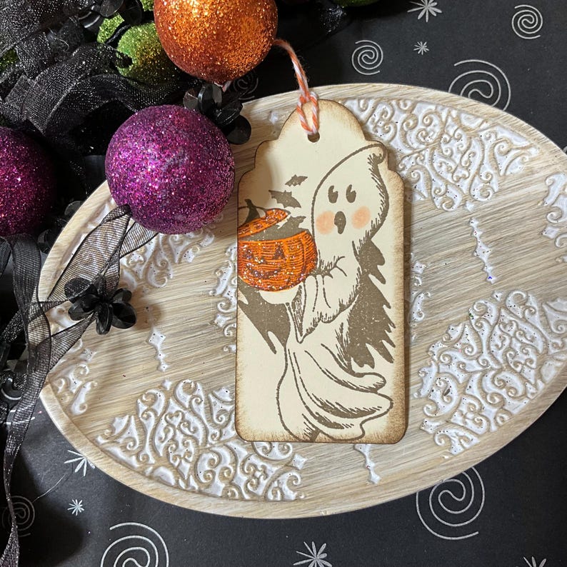 May include: A Halloween-themed tag featuring a cartoon ghost holding an orange jack-o'-lantern. The tag is tan with a scalloped top and a small hole for a string. The ghost has rosy cheeks and is surrounded by bats. The tag is on a decorative wooden tray.