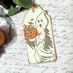 May include: A Halloween-themed gift tag featuring a cartoon ghost holding a jack-o'-lantern. The tag has a scalloped edge and is tied with twine. The ghost has rosy cheeks and is surrounded by bats.
