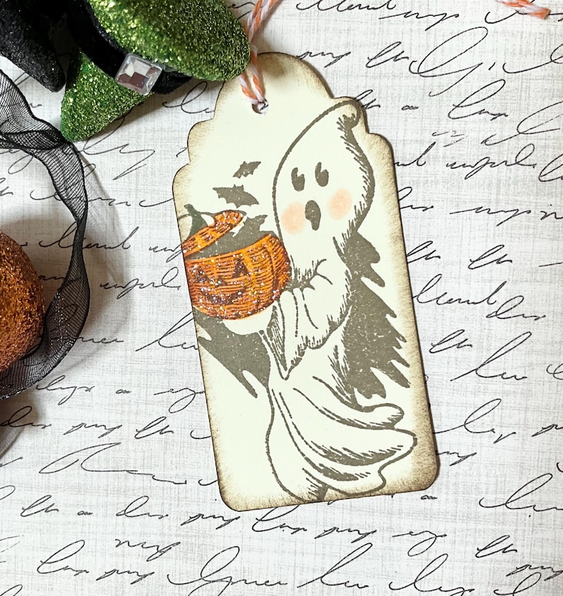 May include: A Halloween-themed gift tag featuring a cartoon ghost holding a jack-o'-lantern. The tag has a scalloped edge and is tied with twine. The ghost has rosy cheeks and is surrounded by bats.