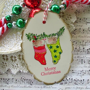 May include: A cream-colored, scalloped-edge Christmas tag with two mice in stockings. One stocking is red, the other green with yellow stars. The tag reads "Merry Christmas" in red. Festive holiday decor.