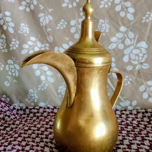 Puede incluir: Una cafetera de metal color latón con base bulbosa, un pico largo y curvado, y una tapa alta y puntiaguda. El recipiente tiene un acabado liso y pulido, con un asa en el lateral y un pequeño emblema circular cerca de la parte superior.
