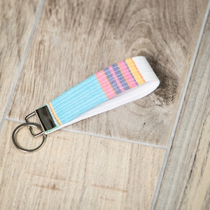 May include: A fabric wristlet key fob with a silver key ring. The key fob features a light blue, pink, and yellow striped pattern on a white background. The key fob is made of woven fabric.