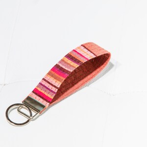 May include: A colorful wristlet keychain with a woven fabric strap. The strap features a pattern of pink, orange, and brown stripes. A silver-colored metal ring is attached for keys.