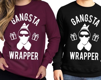 Pode incluir: Duas sweatshirts de manga comprida com um gráfico branco de uma pessoa usando um chapéu de Papai Noel e óculos de sol com o texto "Gangsta Wrapper". A sweatshirt à esquerda é vinho e a sweatshirt à direita é preta.