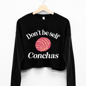 May include: Black long-sleeved cropped sweatshirt with white text that reads "Don't be self Conchas" and a pink and yellow concha pastry graphic.