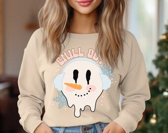 May include: A beige long-sleeve sweatshirt featuring a graphic of a melting snowman wearing light blue earmuffs. The snowman has a carrot nose, pink cheeks, and a dotted smile. Above the snowman, the text CHILL OUT is printed in pink.