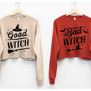 May include: Two cropped sweatshirts with long sleeves. One is beige with the text "Good Witch" and a witch's hat graphic. The other is red with the text "Bad Witch" and a black cat graphic.