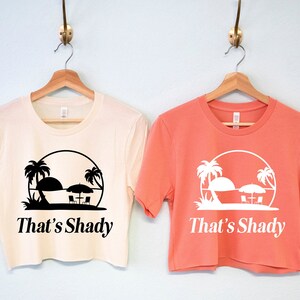 Puede incluir: Dos camisetas recortadas con una escena de playa y el texto "That's Shady". Una camiseta es blanca con texto negro y la otra es coral con texto blanco.