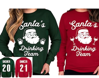 Könnte beinhalten: Zwei Weihnachtspullover mit dem Text "Santa's Drinking Team" und einem Cartoon-Bild von Weihnachtsmann. Ein Pullover ist grün und der andere ist rot. Die Pullover haben weißen Text und Bilder.