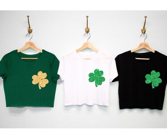 May include: Three crop tops in green, white, and black, each adorned with a glittery shamrock. The green top features a gold shamrock, while the white and black tops have green shamrocks. The shirts are hanging on wooden hangers.