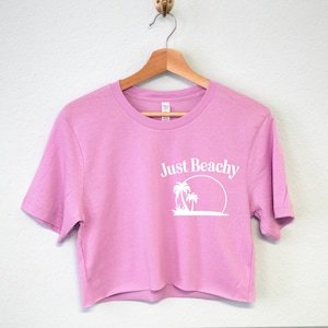 May include: A pink cropped t-shirt with short sleeves and a white graphic of palm trees and a sun with the text "Just Beachy".