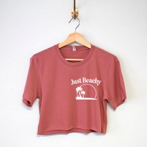 May include: A cropped, short-sleeved t-shirt in a dusty rose color with a white graphic of palm trees and a sun with the text "Just Beachy".