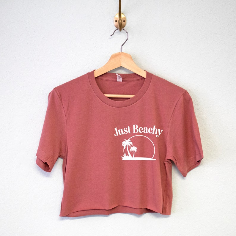 May include: A cropped, short-sleeved t-shirt in a dusty rose color with a white graphic of palm trees and a sun with the text "Just Beachy".