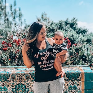 Pu&ograve; includere: Una donna indossa una canotta nera con la scritta "Working off the best lbs I ever gained #babyweight" in lettere rosa, tiene in braccio un bambino che indossa una maglietta a righe bianche e nere con la scritta "Acting like I'm...