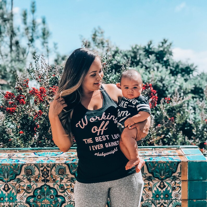 Pu&ograve; includere: Una donna indossa una canotta nera con la scritta "Working off the best lbs I ever gained #babyweight" in lettere rosa, tiene in braccio un bambino che indossa una maglietta a righe bianche e nere con la scritta "Acting like I'm...