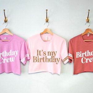 May include: Three pink and red cropped t-shirts with white text. The shirts have white stripes on the sleeves. The text on the shirts reads "Birthday Crew" and "It's my Birthday".