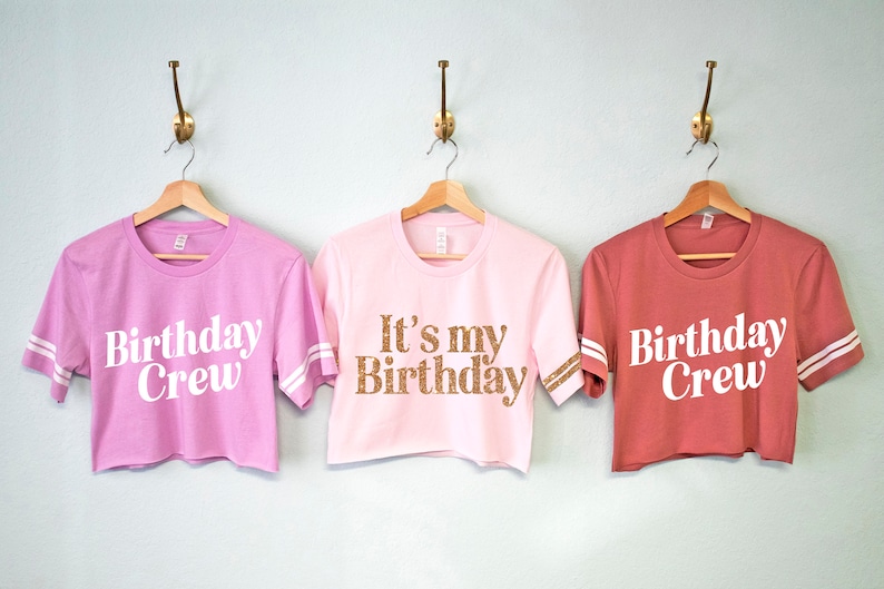 May include: Three pink and red cropped t-shirts with white text. The shirts have white stripes on the sleeves. The text on the shirts reads "Birthday Crew" and "It's my Birthday".