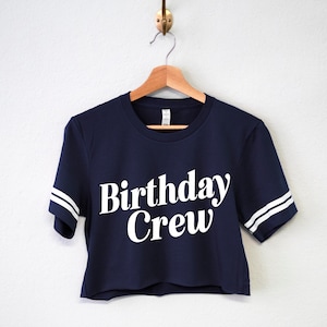 May include: A navy blue cropped t-shirt with white stripes on the sleeves and the text "Birthday Crew" in white.