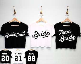Op de afbeelding: Drie zwarte en witte crop tops met de woorden "Bride", "Bridesmaid" en "Team Bride" in een schrijflettertype. De tops hangen aan hangers tegen een witte muur. Onder de tops zijn drie witte shirts met zwarte nummers. De nummers zijn "20", "21" en "88".