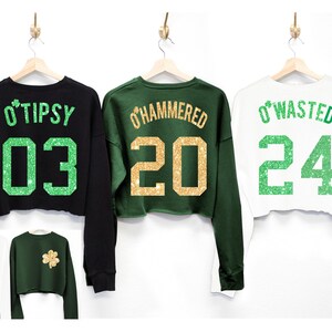 May include: Three cropped sweatshirts with green shamrock designs and gold glitter text. The black sweatshirt says "O'Tipsy 03", the green sweatshirt says "O'Hammered 20", and the white sweatshirt says "O'Wasted 24".