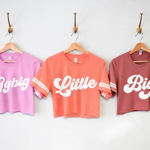 K&ouml;nnte beinhalten: Drei verk&uuml;rzte T-Shirts mit wei&szlig;em Text auf verschiedenen farbigen Shirts. Die Shirts sagen "Ggbig", "Little" und "Big" in einer Retro-Schriftart. Die Shirts haben wei&szlig;e Streifen an den &Auml;rmeln.