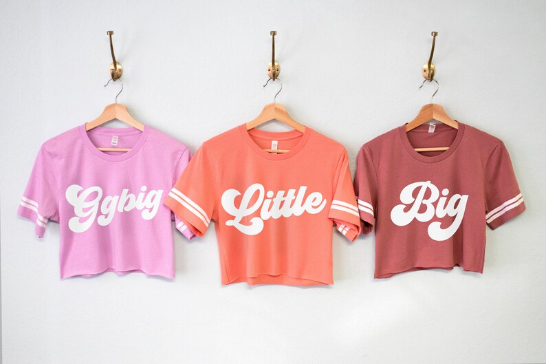 K&ouml;nnte beinhalten: Drei verk&uuml;rzte T-Shirts mit wei&szlig;em Text auf verschiedenen farbigen Shirts. Die Shirts sagen "Ggbig", "Little" und "Big" in einer Retro-Schriftart. Die Shirts haben wei&szlig;e Streifen an den &Auml;rmeln.