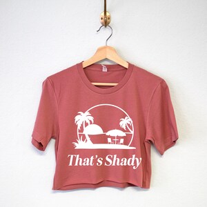Puede incluir: Una camiseta recortada de manga corta en color rosa polvoriento. La camiseta presenta un gr&aacute;fico blanco de palmeras, una sombrilla y el texto "That's Shady".
