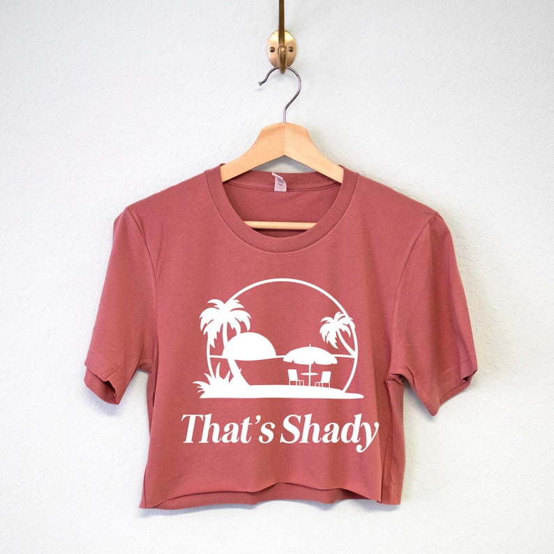Puede incluir: Una camiseta recortada de manga corta en color rosa polvoriento. La camiseta presenta un gr&aacute;fico blanco de palmeras, una sombrilla y el texto "That's Shady".