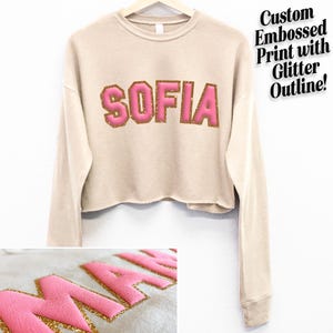 May include: A beige cropped sweatshirt with long sleeves features the name "SOFIA" in pink letters with a gold glitter outline. The sweatshirt is made of a soft, comfortable material.