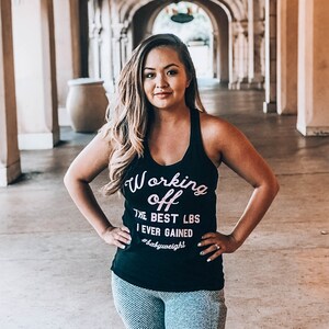 Pu&ograve; includere: Una donna indossa una canotta nera con la scritta "Working off the best lbs I ever gained #babyweight" in lettere rosa. &Egrave; in piedi in un corridoio con archi.