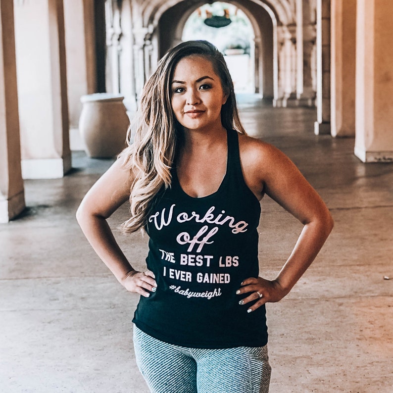 Pu&ograve; includere: Una donna indossa una canotta nera con la scritta "Working off the best lbs I ever gained #babyweight" in lettere rosa. &Egrave; in piedi in un corridoio con archi.