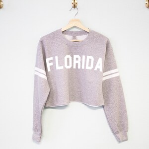 以下が含まれることがあります： 白い袖のストライプと前面に白い「FLORIDA」の文字がプリントされたグレーのクロップドスウェットシャツです。