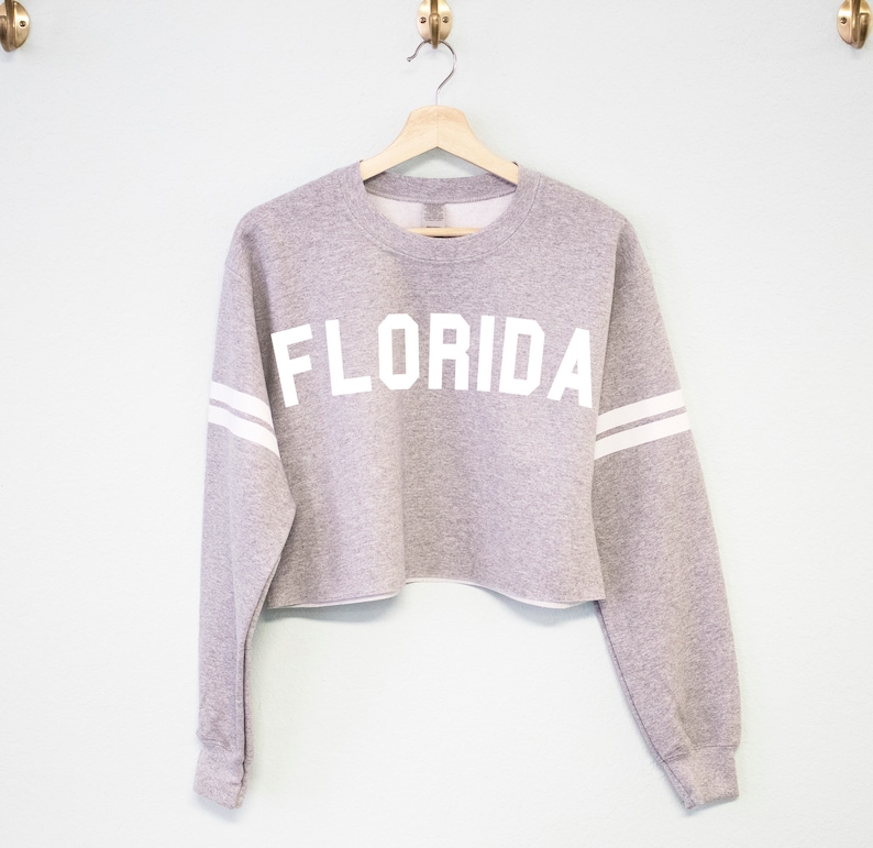 以下が含まれることがあります： 白い袖のストライプと前面に白い「FLORIDA」の文字がプリントされたグレーのクロップドスウェットシャツです。