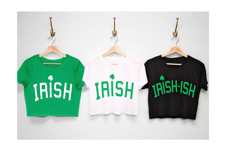 May include: Three cropped t-shirts hanging on hangers. The shirts are green, white, and black. Each shirt has a green shamrock and the word "IRISH" in white letters. The black shirt says "IRISH-ISH".