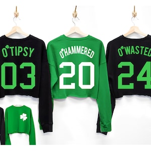 May include: Three black and green long-sleeved cropped sweatshirts with shamrock designs and numbers on the back. The sweatshirts say "O'Tipsy 03", "O'Hammered 20", and "O'Wasted 24".