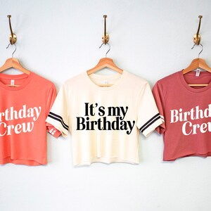 May include: Three cropped t-shirts with white text on different colored shirts. The shirts have black and white stripes on the sleeves. The text on the shirts reads "Birthday Crew", "It's my Birthday", and "Birthday Crew".