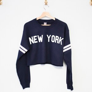 以下が含まれることがあります： 白の長い袖と「NEW YORK」という白い文字が書かれたネイビーブルーのクロップドスウェットシャツです。