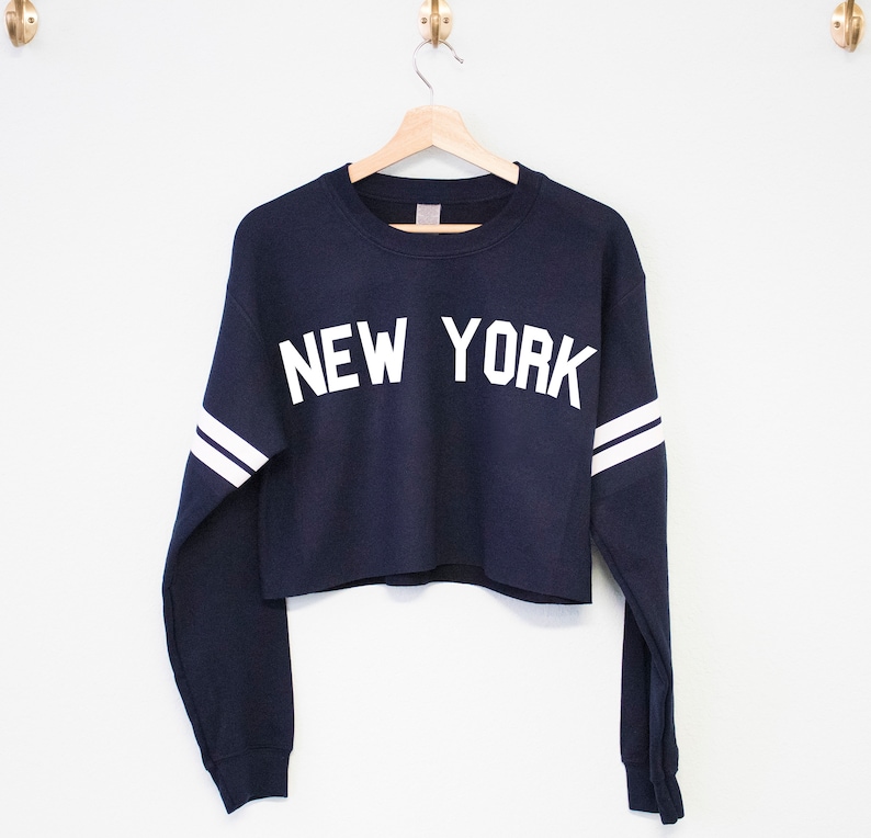 以下が含まれることがあります： 白の長い袖と「NEW YORK」という白い文字が書かれたネイビーブルーのクロップドスウェットシャツです。