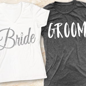 Puede incluir: Dos camisetas, una blanca y otra gris, con las palabras "Bride" y "Groom" impresas en ellas. La camiseta de la novia tiene un dise&ntilde;o brillante.