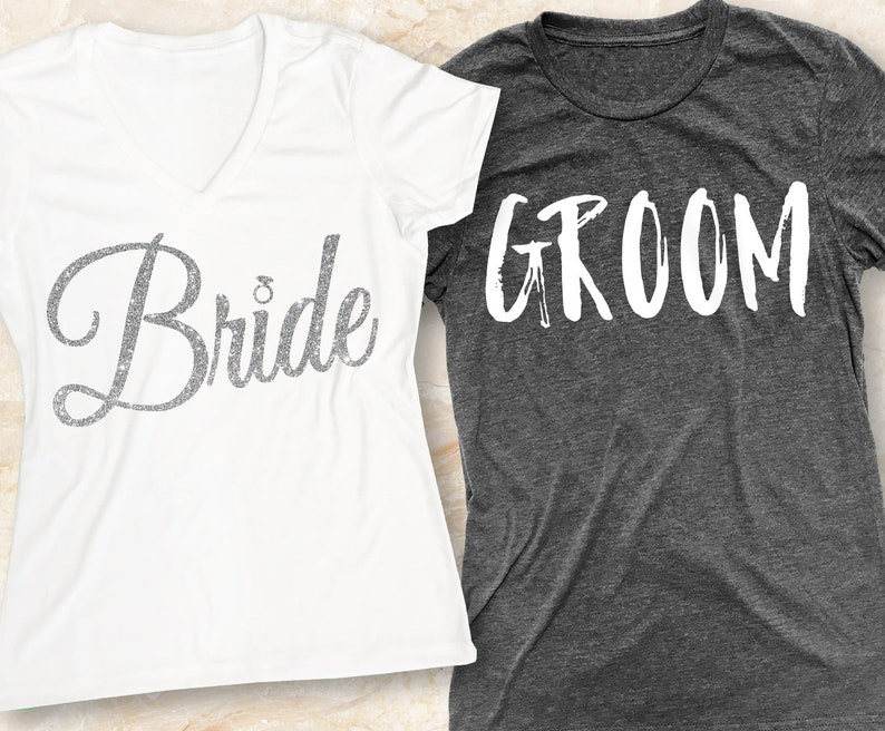 Puede incluir: Dos camisetas, una blanca y otra gris, con las palabras "Bride" y "Groom" impresas en ellas. La camiseta de la novia tiene un dise&ntilde;o brillante.