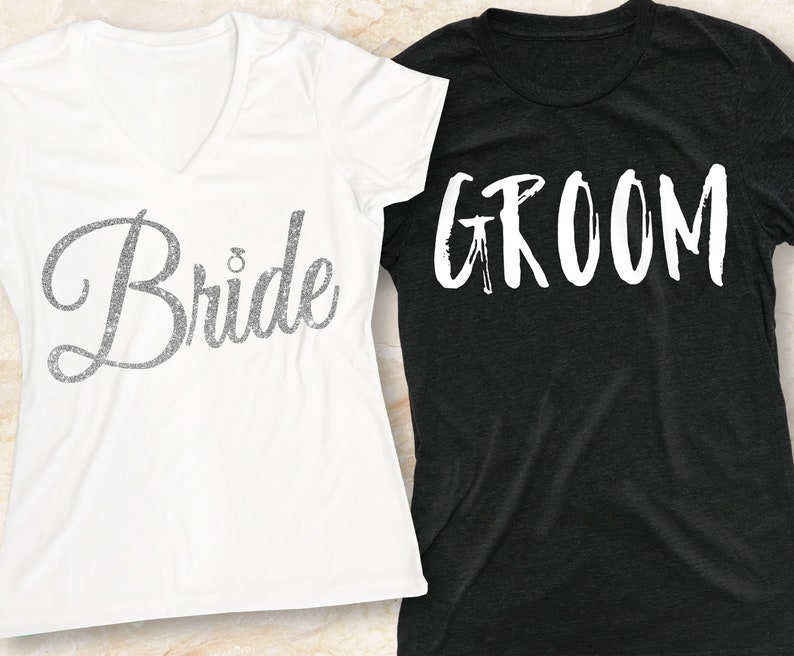 Puede incluir: Dos camisetas blancas con las palabras "Bride" y "Groom" impresas en ellas. La camiseta "Bride" tiene un dise&ntilde;o de purpurina plateada. La camiseta "Groom" tiene un fondo negro.