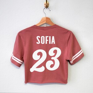K&ouml;nnte beinhalten: Ein rotes, kurzes T-Shirt mit wei&szlig;en Buchstaben auf der R&uuml;ckseite. Das Shirt tr&auml;gt die Aufschrift "SOFIA 23" in einer wei&szlig;en, stilisierten Schrift.