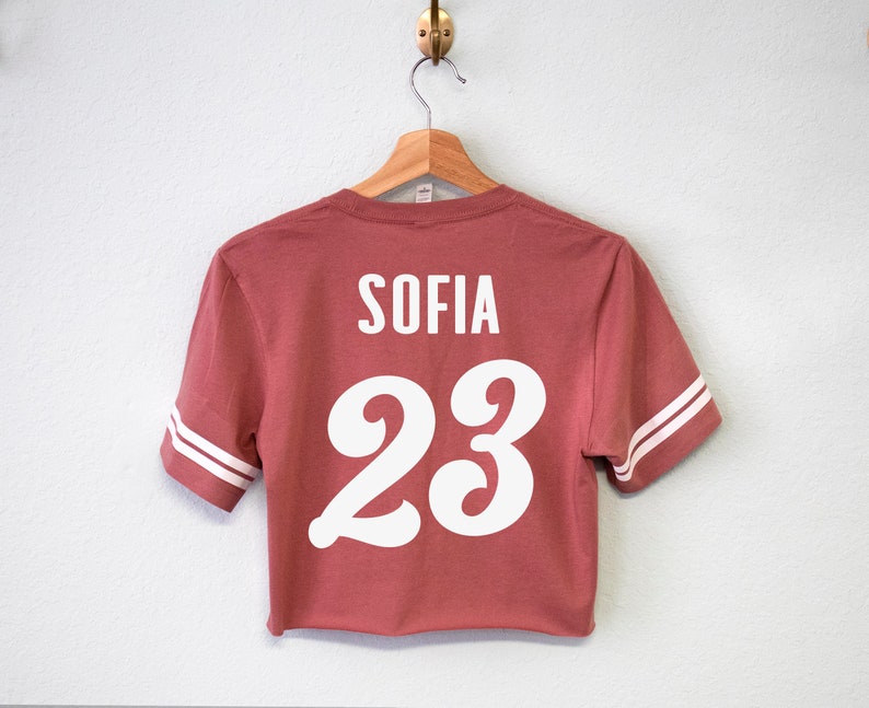 K&ouml;nnte beinhalten: Ein rotes, kurzes T-Shirt mit wei&szlig;en Buchstaben auf der R&uuml;ckseite. Das Shirt tr&auml;gt die Aufschrift "SOFIA 23" in einer wei&szlig;en, stilisierten Schrift.