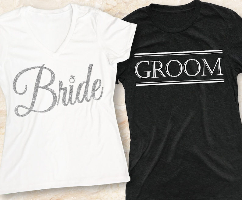 Puede incluir: Dos camisetas blancas con las palabras "Bride" y "Groom" impresas en ellas. La camiseta "Bride" tiene un dise&ntilde;o de purpurina plateada. La camiseta "Groom" tiene un borde de doble l&iacute;nea alrededor de la palabra.