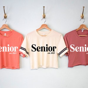 May include: Three cropped t-shirts with the word "Senior" and the text "est. 2023" on them. The shirts are in coral, cream, and dusty rose.